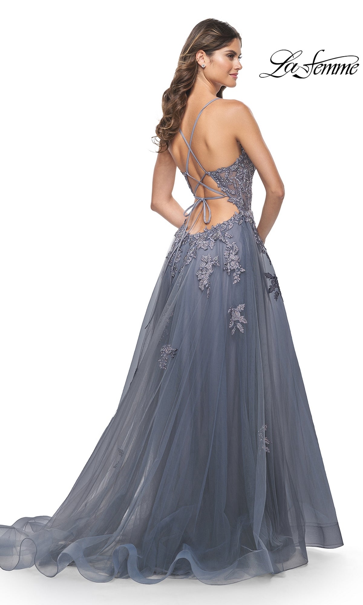 A woman stands against a white background wearing the La Femme Plunging V-Neck Long Prom Ball Gown 31472, featuring a slate blue hue, floral lace details, an open back with crisscross laces, and a flowing formal silhouette.